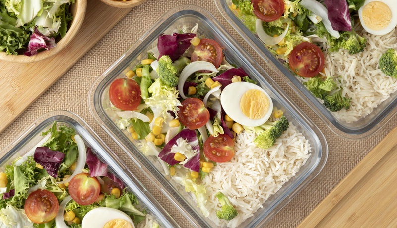 Prepared salads with vegetables and rice in meal prep containers on a wooden surface.